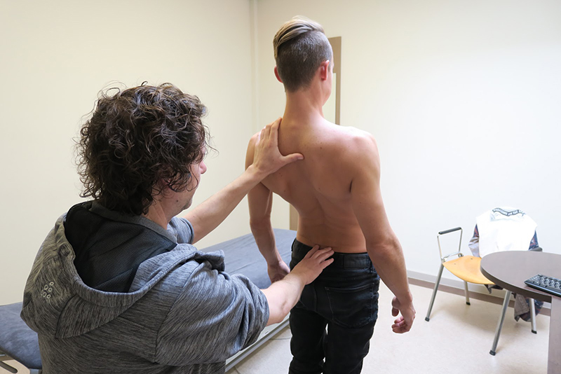 Manueel therapeut Tim met een grijze hoodie helpt een patiënt zonder shirt aan zijn rugklachten in de behandelkamer.